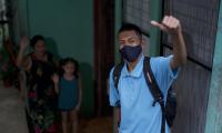 Estudiante con mascarilla saliendo de su casa. Su familia se ve al fondo despidiéndose.